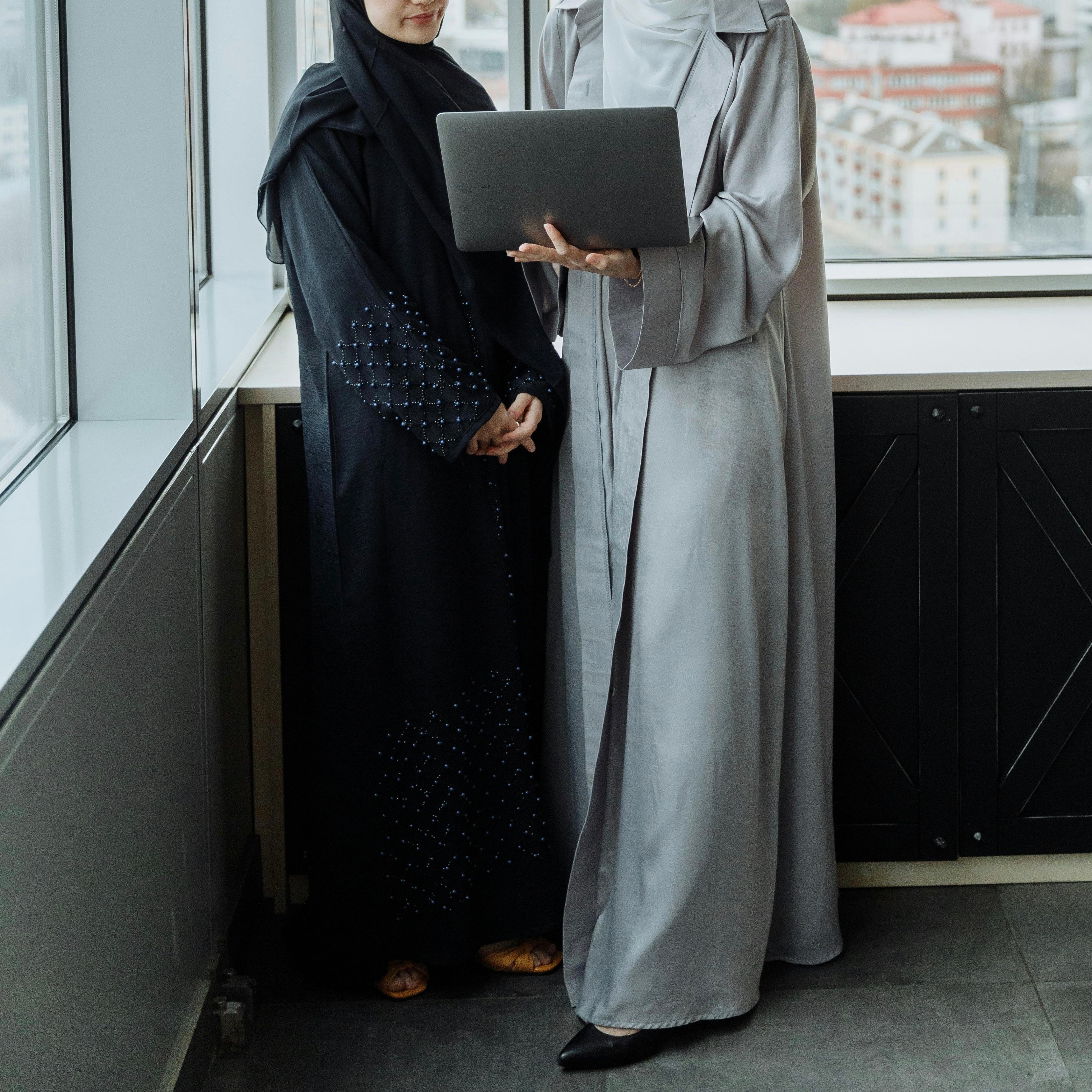 Two women in abayas standing by a window in a modern office, one in a black embellished abaya and the other in a light grey abaya, looking at a laptop together.
