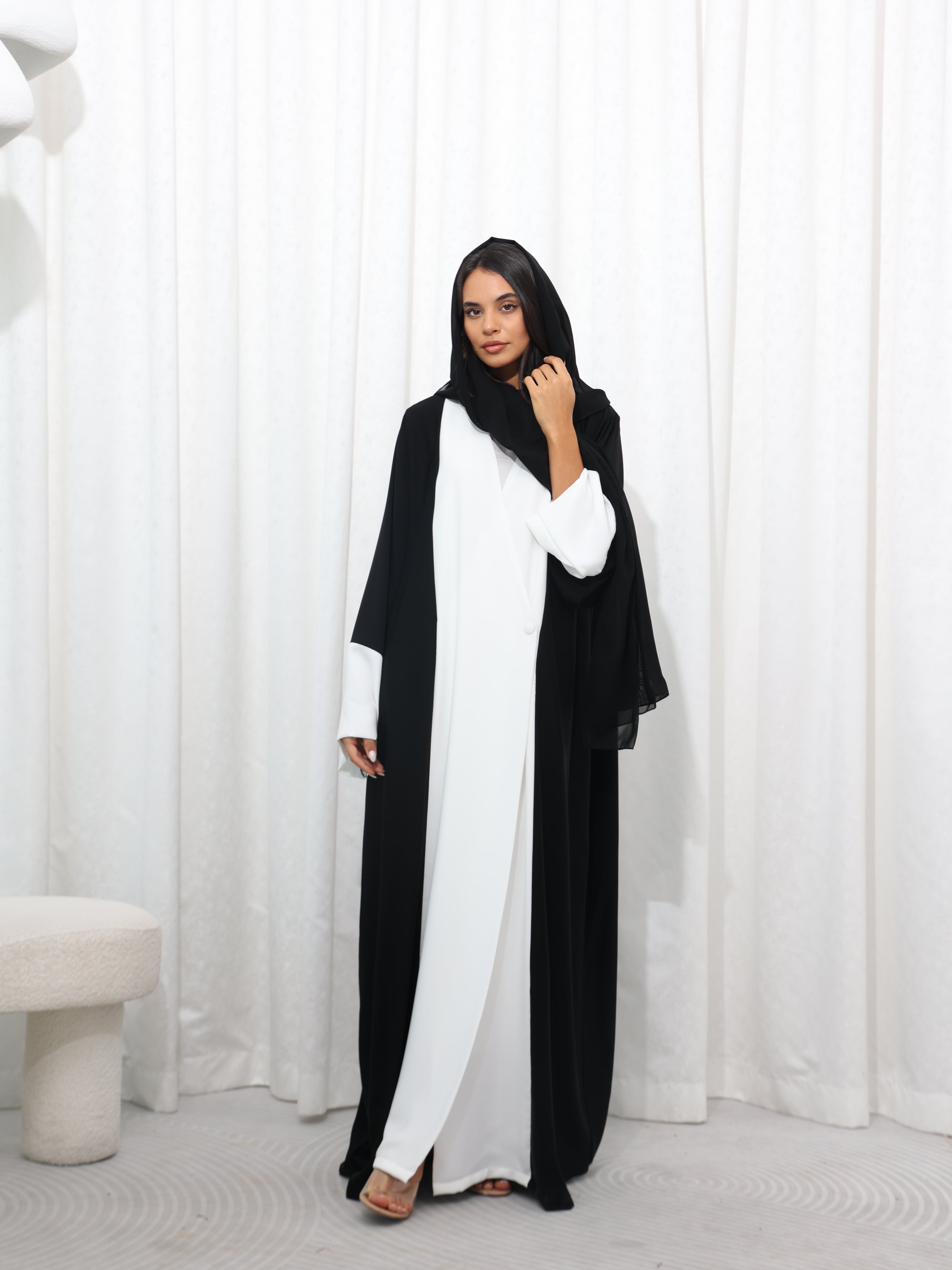 Model wearing black and white RUMI abaya with black sheila , standing in front of a white curtain backdrop