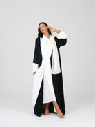 Model posing with one arm raised in RUMI black and white tux like abaya, styled with white scarf and open front