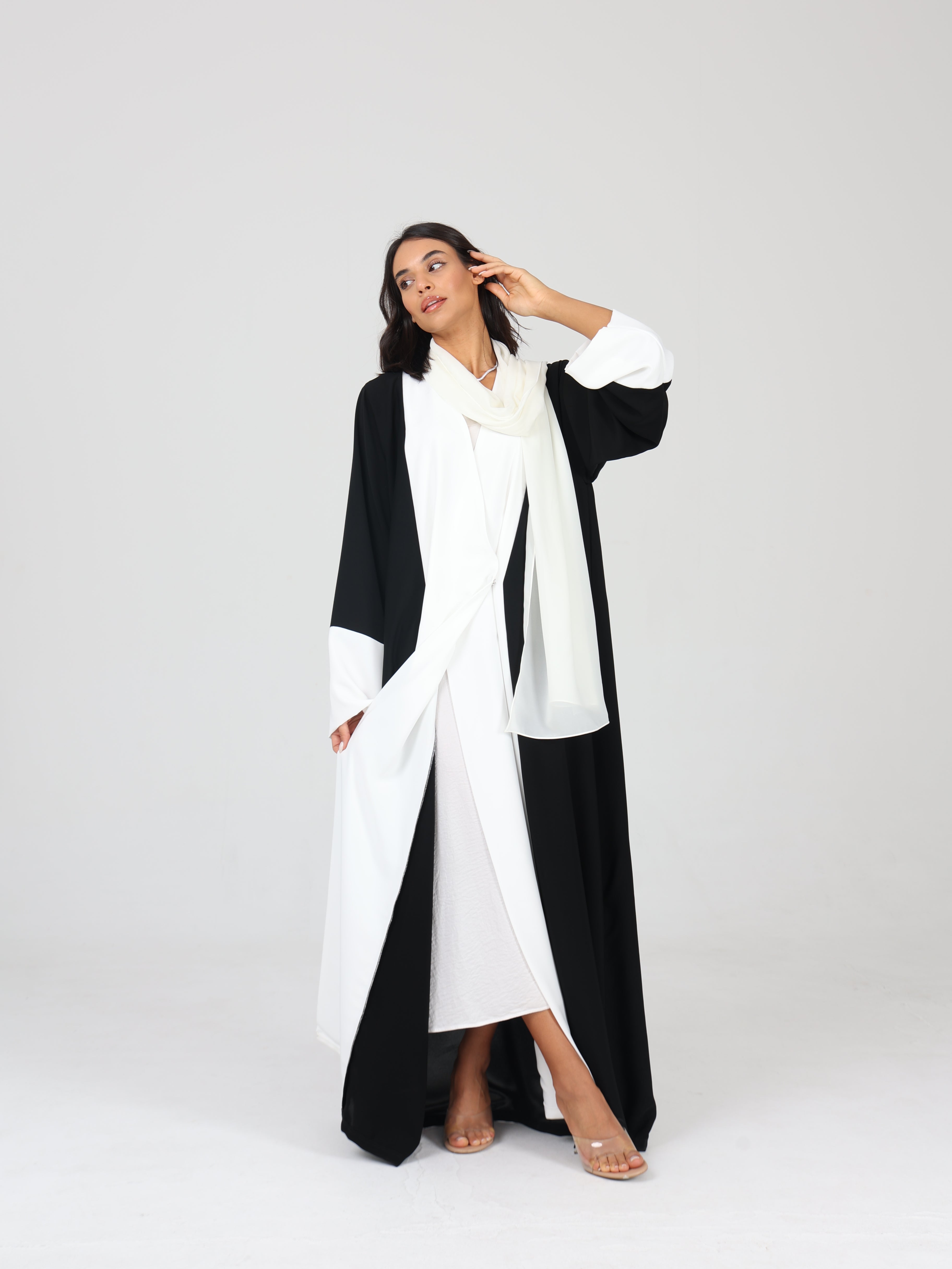 Model posing with one arm raised in RUMI black and white tux like abaya, styled with white scarf and open front
